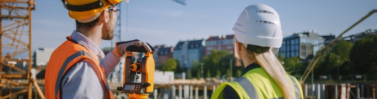 Surveyors wearing high-visibility vests and safety helmets while using a yellow surveying instrument on a construction site